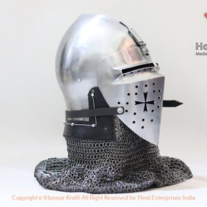 May include: A silver helmet with a visor and a chainmail coif. The helmet has a cross on the visor and is decorated with small holes. The helmet is on a white background.