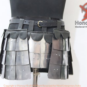 Puede incluir: Una falda de metal plateado con un cinturón de cuero negro y un borde festoneado. La falda está hecha de placas superpuestas y está asegurada con remaches. La falda se muestra en un maniquí.