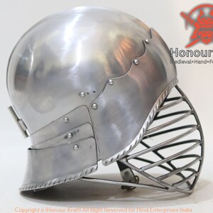 Op de afbeelding: Een zilveren helm met een vizier en een decoratieve rand. De helm is gemaakt van metaal en heeft een afgeronde vorm. Het vizier is gemaakt van metalen staven en is met scharnieren aan de helm bevestigd.