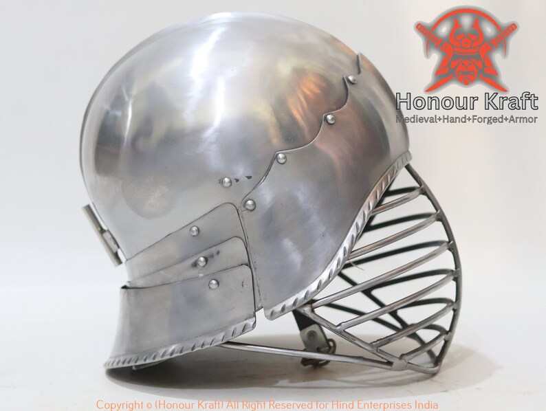 Op de afbeelding: Een zilveren helm met een vizier en een decoratieve rand. De helm is gemaakt van metaal en heeft een afgeronde vorm. Het vizier is gemaakt van metalen staven en is met scharnieren aan de helm bevestigd.