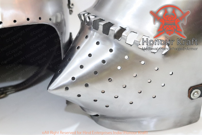 K&ouml;nnte beinhalten: Zwei mittelalterliche Helme, einer mit Visier und der andere mit geschlossenem Gesicht, beide aus Metall mit silberner Oberfl&auml;che und kleinen L&ouml;chern. Der Helm mit dem Visier hat einen spitzen Nasenschutz. Der Text "Honour Kraft Medieval Hand Forged Armor" ist auf dem Helm mit dem Visier sichtbar.