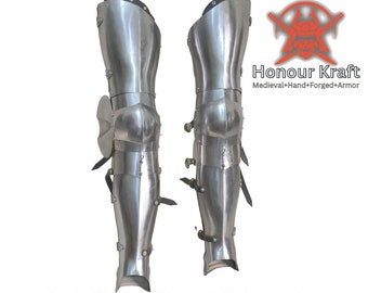 May include: A pair of silver-colored steel medieval leg armor, also known as greaves, with knee pads and straps. The armor is hand-forged and is made for reenactment or display.