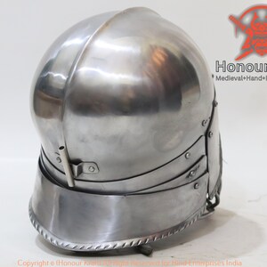Op de afbeelding: Een zilveren helm met een afgeronde bovenkant en een vizier. De helm heeft een decoratieve rand rond de onderkant. De helm is gemaakt van metaal en heeft een glanzende afwerking.