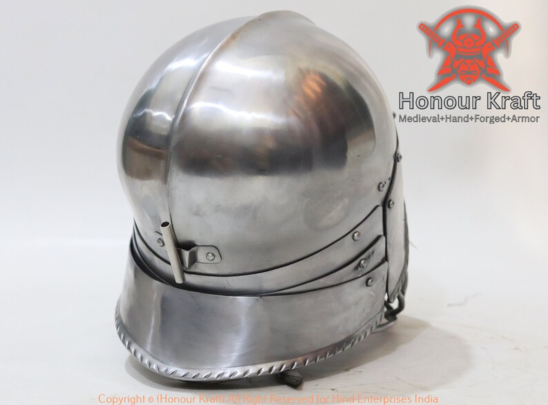 Op de afbeelding: Een zilveren helm met een afgeronde bovenkant en een vizier. De helm heeft een decoratieve rand rond de onderkant. De helm is gemaakt van metaal en heeft een glanzende afwerking.