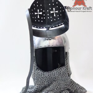 May include: A silver helmet with a black visor and a chainmail coif. The helmet has a cross design on the visor and the text "Honour Kraft" is visible on the right side of the image.