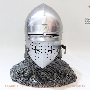 May include: A silver helmet with a visor and a chainmail coif. The helmet has a cross design on the visor and is decorated with small rivets. The helmet is on a white background.