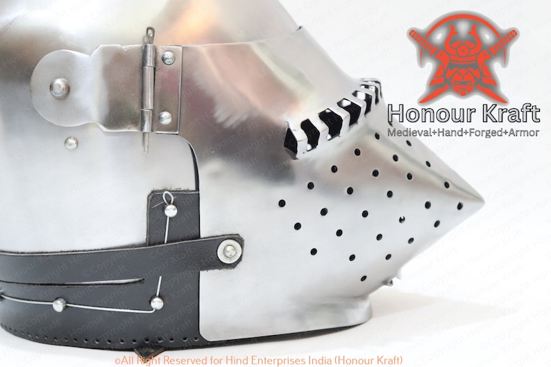 K&ouml;nnte beinhalten: Nahaufnahme eines silbernen Helms mit einem spitzen Visier und einem schwarzen Lederriemen. Der Helm hat eine Reihe kleiner L&ouml;cher am Visier und ein dekoratives Muster auf der Oberseite. Der Text "Honour Kraft" ist im Bild sichtbar.