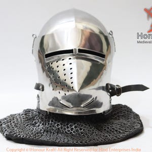 May include: A silver helmet with a visor and a chainmail coif. The helmet has a pointed nose guard and a series of small holes for ventilation. The helmet is sitting on a white background.