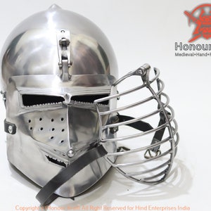 Puede incluir: Un casco de plata con una máscara facial articulada. El casco tiene una parte superior puntiaguda y una visera con pequeños agujeros. La máscara facial está hecha de barras de metal y está articulada en el lateral. El casco es un casco de estilo medieval.