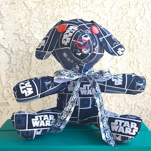 May include: A plush toy shaped like a dog with a black and white Star Wars print. The toy has red buttons for eyes and a white ribbon around its neck.