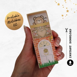 May include: A printable matchbox kit featuring a sleeping bear illustration. The matchbox design includes a brick facade, a white door with a gold knocker, and a heart-shaped design. The text "Printable Matchbox Kit" is on a gold circle.