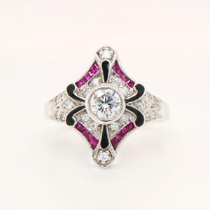 May include: A silver ring with a central, round, clear gemstone. The ring features a geometric design with black enamel accents, small clear gemstones, and vibrant pink gemstones. The ring is set against a white background.