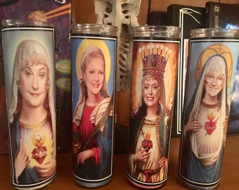 May include: Four glass prayer candles featuring portraits of women in religious iconography. Each candle has a different portrait, with a halo and a flaming heart. The candles are cylindrical and have a black rim.
