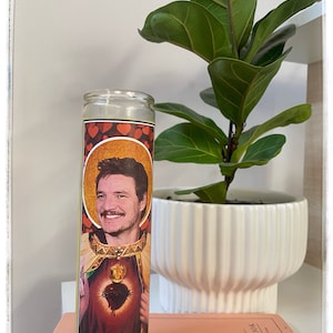 Può includere: Una candela di preghiera in vetro con un ritratto di un uomo con i baffi, circondato da un'aureola dorata. La candela ha un motivo a cuori rossi nella parte superiore e un'immagine del Sacro Cuore. Sullo sfondo c'è una pianta in vaso bianca.