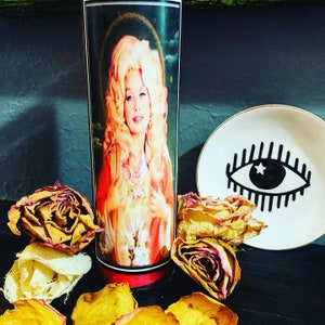 Puede incluir: Una vela alta con un retrato de una mujer con cabello rubio, vestida con un atuendo rojo y dorado. Un plato blanco pequeño con un diseño de ojo negro está al lado de la vela. Pétalos y rosas secas están esparcidos alrededor de la vela.