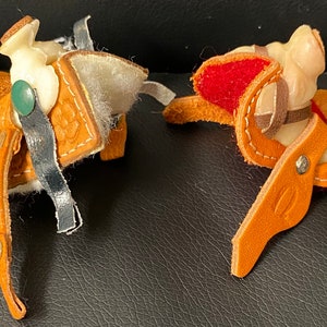 May include: Two miniature leather saddles with keychains. One saddle is brown with a green and white fringe and the other is brown with a red and white fringe.