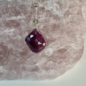 May include: A faceted, deep magenta-colored gemstone pendant suspended from a silver chain. The pendant features a small, translucent, pale green bead accent above the main stone, connected by silver wire wrapping to the metal bail.