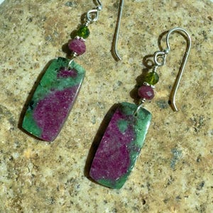 May include: A pair of dangle earrings featuring rectangular ruby and zoisite stone pendants. Each earring includes a small faceted green bead and a faceted pink bead attached to a silver-toned metal hook wire for pierced ears.