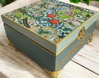 Puede incluir: Una caja de madera cuadrada y decorativa con un diseño floral en la tapa. La caja está pintada en un color verde azulado apagado con detalles dorados en las esquinas y los bordes. La tapa presenta un patrón colorido de flores y hojas.