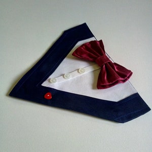 May include: A white fabric dog collar with a navy blue trim and a burgundy bow tie. The collar has four white buttons and a red heart-shaped button.