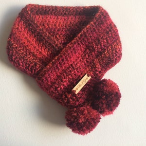 Könnte beinhalten: Roter und schwarzer Häkel-Halswärmer mit zwei flauschigen Bommeln. Der Halswärmer hat ein Holzschild mit der Aufschrift "Elle & Grey".
