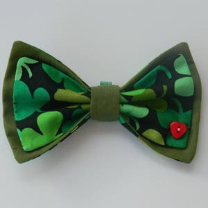 May include: A handmade bow tie with a green shamrock pattern on a black background. The bow tie has a dark green border and a small red heart-shaped button. The bow tie is a festive accessory.