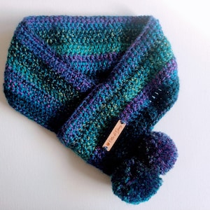 May include: A blue, green and purple crocheted infinity scarf with two pom-poms and a brown leather tag that says "Ellie & Gray".