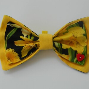 May include: A yellow bow tie with a black background and a floral pattern of yellow daffodils. The bow tie has a red heart-shaped button in the center.