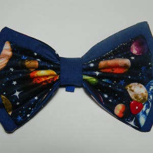 Puede incluir: Una pajarita azul con un estampado de temática espacial con planetas, estrellas y un botón rojo.