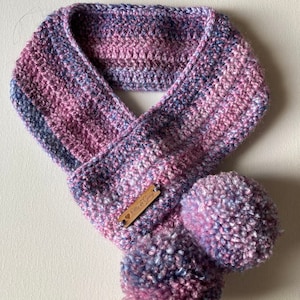 May include: A purple and blue crocheted scarf with two pom-poms and a wooden tag that says "Ella & Henry".