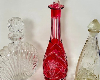May include: Three glass decanters with stoppers. The decanter on the left is clear glass with a scalloped design. The decanter in the center is red glass with a cut glass design. The decanter on the right is clear glass with a geometric design.