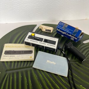 May include: A vintage Minolta Pocket Autopak 70 camera with accessories. Includes a camera, owner's manual, film cartridges, a flash cube box, and a camera strap. The camera is black and white with a yellow button.