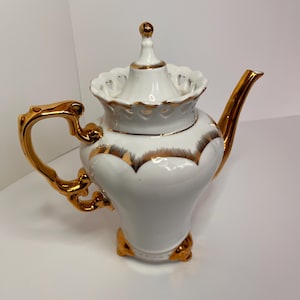 Puede incluir: Tetera de porcelana blanca con detalles dorados. La tetera tiene una tapa con un diseño decorativo y un asa dorada. La tetera está sobre una superficie blanca.