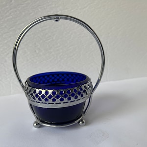 Puede incluir: Un azucarero de vidrio azul cobalto con un asa y una base de alambre plateado. El tazón tiene un diseño decorativo de alambre plateado alrededor del borde.