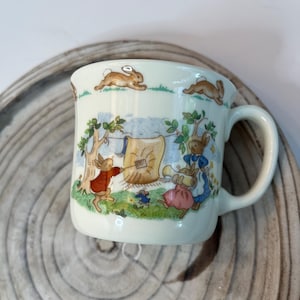 Può includere: Tazza in ceramica color crema con un'illustrazione stravagante di conigli e altri animali in una scena pastorale. Il design include uno stendibiancheria con oggetti appesi, alberi e conigli in corsa. La tazza ha un manico curvo e un bordo leggermente svasato.