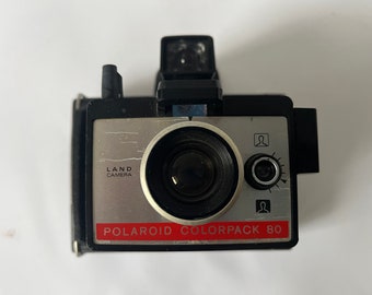 May include: A black and silver Polaroid Colorpack 80 instant camera with a red label. The camera has a lens, a flash, and a viewfinder.