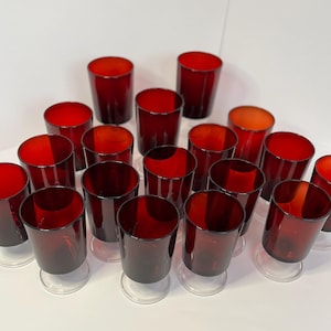Könnte beinhalten: Eine Sammlung von 18 roten Glas-Trinkgläsern mit klaren Böden. Die Gläser sind in einem Halbkreis auf einer weißen Oberfläche angeordnet.