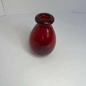 Könnte beinhalten: Eine kleine, rote Glasvase mit abgerundetem Boden und einer leicht ausgestellten Öffnung.