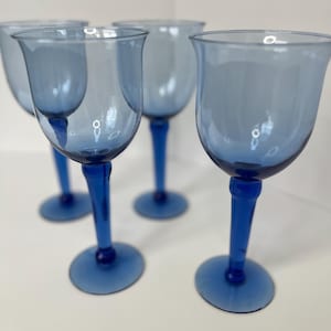 Op de afbeelding: Set van vier blauwe glazen wijnglazen met een heldere kom en een blauwe steel. De glazen zijn in een rij op een wit oppervlak gerangschikt.