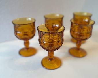 May include: Set of five amber glass stemmed goblets with a textured design. The goblets are arranged in a semi-circle on a white surface.