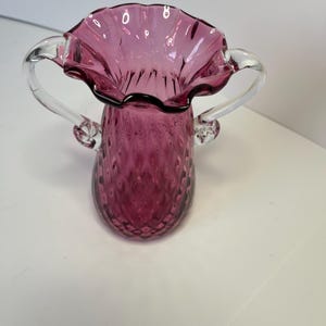 May include: A small, pink glass vase with clear glass handles. The vase has a ruffled top and a textured, diamond-shaped pattern.