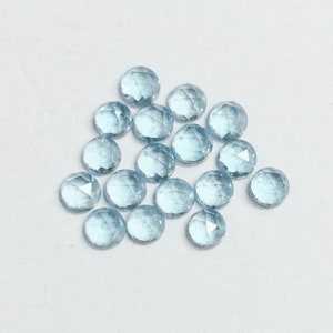 May include: A collection of light blue round faceted gemstones. The gemstones are scattered on a white surface.