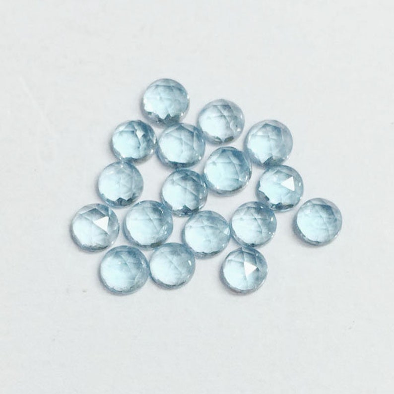 May include: A collection of light blue round faceted gemstones. The gemstones are scattered on a white surface.