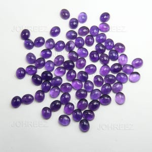 May include: A collection of oval-shaped, polished amethyst gemstones in varying shades of purple. The gemstones are scattered across a white surface, showcasing their smooth, reflective surfaces. The gemstones are ideal for jewelry making.