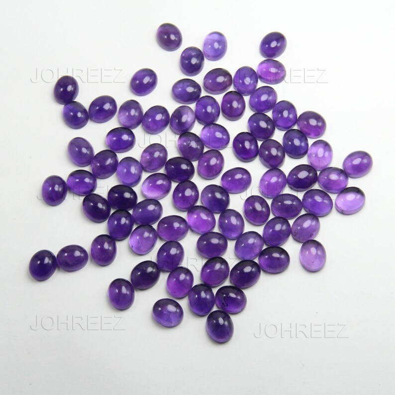 May include: A collection of oval-shaped, polished amethyst gemstones in varying shades of purple. The gemstones are scattered across a white surface, showcasing their smooth, reflective surfaces. The gemstones are ideal for jewelry making.
