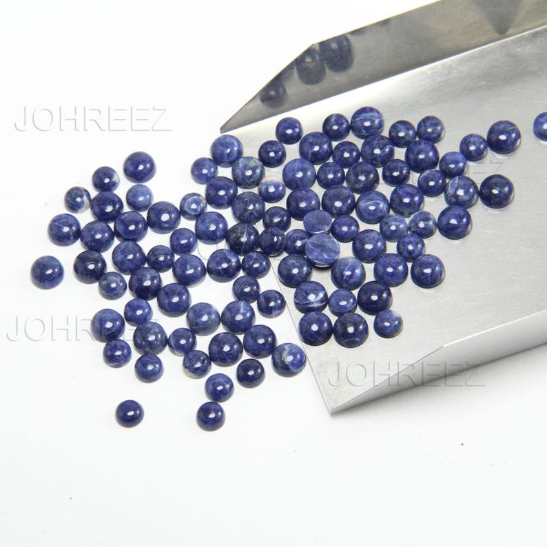 May include: A large group of blue gemstone cabochons, likely sodalite, in various sizes, scattered on a silver tray. The cabochons are smooth and polished, with a slightly iridescent sheen.