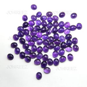 May include: A collection of oval-shaped, polished amethyst gemstones in varying shades of purple. The gemstones are scattered on a white surface, showcasing their smooth, reflective surfaces. The image is well-lit, highlighting the color and clarity of the stones.
