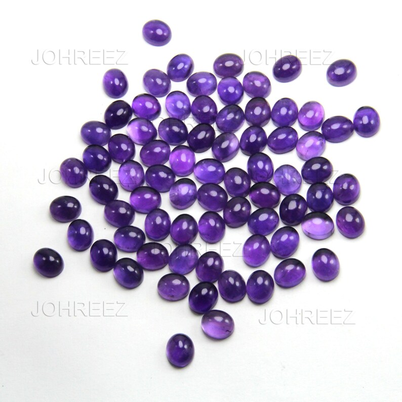 May include: A collection of oval-shaped, polished amethyst gemstones in varying shades of purple. The gemstones are scattered on a white surface, showcasing their smooth, reflective surfaces. The image is well-lit, highlighting the color and clarity of the stones.