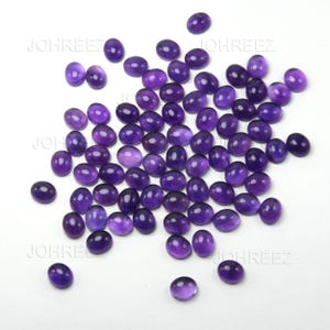 May include: A collection of oval-shaped, polished amethyst gemstones. The gemstones are a deep purple color and are scattered across a white surface. The gemstones are smooth and have a glossy finish. The image is well-lit, highlighting the color and clarity of the stones.