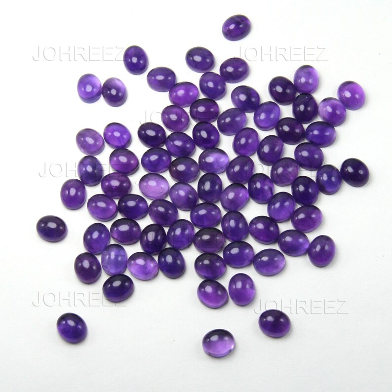 May include: A collection of oval-shaped, polished amethyst gemstones. The gemstones are a deep purple color and are scattered across a white surface. The gemstones are smooth and have a glossy finish. The image is well-lit, highlighting the color and clarity of the stones.